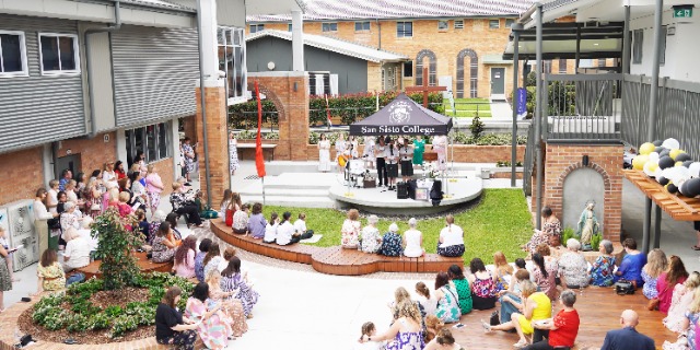 school community gathered around a courtyard stage with students presenting, highlighting school events and shared learning experiences